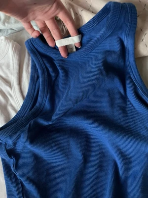 Ribbade linnetoppar från H&M - Två ribbade linnetoppar från H&M i blått och vitt. Båda är ärmlösa och har en klassisk rund halsringning. Perfekta att styla till jeans eller kjol för en enkel och snygg look.