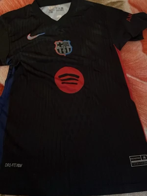 Svart FC Barcelona matchtröja Nike - Snygg svart FC Barcelona matchtröja från Nike med klubbmärke, röd sponsorlogga och Dri-FIT ADV-tyg. På ryggen står det Lamine Yamal och nummer 19 i vitt, samt UNHCR-logga. Perfekt för dig som älskar fotboll och vill ha en unik supportertröja.