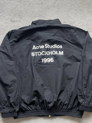 Acne Studios Svart vindjacka med logo baksida - Svart vindjacka med klassisk krage och hel dragkedja framtill. Jackan har resår vid ärmslut och midja för en skön passform. Tillverkad i lätt och vindtätt material, perfekt för blåsiga dagar. Enkel och stilren design utan synliga loggor.
