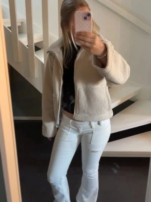 Vita flare jeans med slitning - Säljer ett par vita flare jeans med hög midja och snygg slitning vid låret. Jeansen har dubbla knappar i midjan och klassiska fickor både fram och bak. Materialet är stretchigt och bekvämt, perfekt för en trendig look.