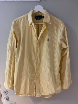 Gulvit randig skjorta från Ralph Lauren - Klassisk skjorta från Ralph Lauren i gulvit smalrandig bomull. Långärmad modell med button-down krage och broderad blå logga på bröstet. Perfekt för en clean och stilren look. Priset är ej hugget i sten vid snabb affär
