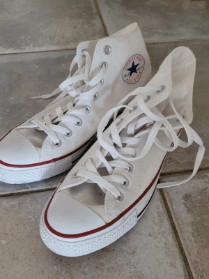 Vita Converse All Star high tops - Klassiska vita Converse All Star high top sneakers med röd rand runt sulan och den ikoniska loggan på sidan. Skorna har vita snören, metallöljetter och är tillverkade i canvas med gummisula. Perfekta för en avslappnad och cool stil.