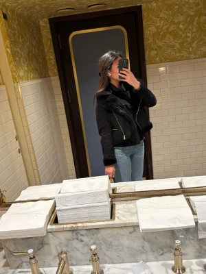 Svart skinnjacka med fuskpälsdetaljer - Snygg svart skinnjacka med fuskpälsdetaljer på kragen och längs dragkedjan. Jackan har en klassisk bikerstil med snedställd dragkedja och fickor framtill. Perfekt för dig som vill ha en trendig och tidlös look.