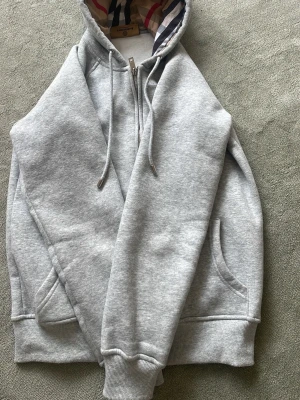 Grå hoodie från Burberry med huva - Snygg grå hoodie från Burberry med dragkedja och klassiskt rutigt foder i huvan. Hoodien har fickor på sidorna, ribbade muddar och justerbar dragsko i huvan. Perfekt för dig som vill ha en stilren och exklusiv look med ikoniska Burberry-detaljer.