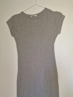 Grå klänning  - En enkel och stilren grå figurnära klänning från Pull&Bear. Klänningen har rund halsringning och korta ärmar. Tillverkad i mjuk bomull som känns skön mot huden. Perfekt för en avslappnad look.