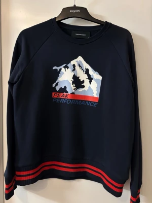 Mörkblå sweatshirt Peak Performance - Cool mörkblå sweatshirt från Peak Performance med bergmotiv på bröstet och texten 'Peak Performance'. Tröjan har ribbade muddar med röda och blå ränder vid ärmslut och nederkant. Perfekt för dig som gillar sportig stil.