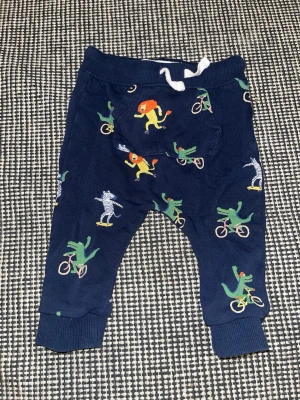 Blå mjukisbyxor med djurmönster - Mörkblå mjukisbyxor med färgglada tryck av lejon, krokodiler och zebror som åker skateboard och cyklar. Byxorna har mudd vid benslut, resår i midjan med vit dragsko och en liten ficka framtill. Perfekta för en lekfull och avslappnad stil.