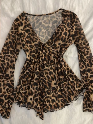 Leopardmönstrad mesh topp med knyt - Säljer en transparent leopardmönstrad topp i mesh med v-ringning och knyt framtill. Toppen har långa ärmar och en lätt utsvängd passform som ger en snygg siluett. Perfekt för dig som vill sticka ut lite extra.