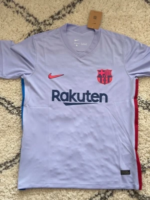 Ljuslila FC Barcelona matchtröja Nike - Snygg ljuslila FC Barcelona matchtröja från Nike med klubbmärke och Rakuten-tryck på bröstet. Röda och blå detaljer på sidorna, korta ärmar och V-ringning. Tillverkad i lätt och ventilerande material, perfekt för fotboll eller träning.