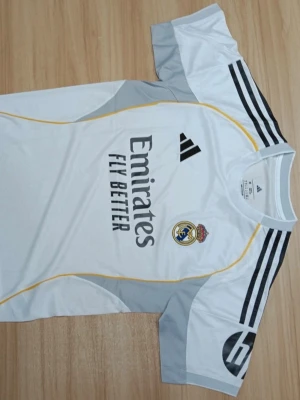 Real Madrid matchtröja Adidas vit - Snygg vit Real Madrid matchtröja från Adidas med svarta detaljer och gula linjer. Tröjan har klubbmärke, sponsortryck 'Emirates Fly Better' och klassiska Adidas-ränder på axlarna. Tillverkad i lätt och ventilerande material, perfekt för fotbollsträning eller match.