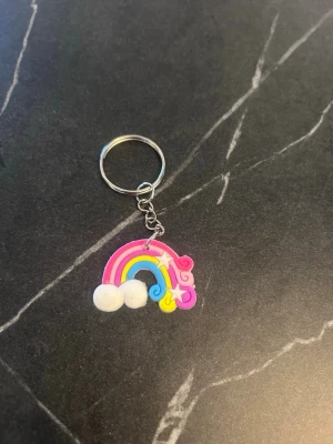 Nyckelring med regnbåge och moln - Färgglad nyckelring med en regnbåge i rosa, gul och blå samt vita moln och en liten stjärna. Tillverkad i plast och metall, perfekt att hänga på väskan eller nyckelknippan för en lekfull touch.