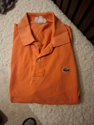 Orange pikétröja från Lacoste - Klassisk orange pikétröja från Lacoste med den ikoniska krokodilloggan på bröstet. Tröjan har krage, knappar framtill och är tillverkad i ett luftigt bomullsmaterial. Perfekt för en avslappnad och stilren look.