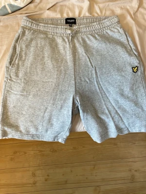 Grå bomullsshorts från Lyle & Scott - Säljer ett par ljusgrå shorts från Lyle & Scott med elastisk midja och snörning. Shortsen har sidofickor och en bakficka, samt den klassiska Lyle & Scott-loggan på vänster ben. Perfekta för sommarens chill och sport. Dom är inprincip nya men när jag kollade lite noggrannare på dem så såg jag att det var två små hål som syns i de två sista bilderna. Det syns inte om man inte kollar noga och går säkert att fixa lätt.