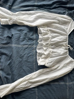 Vit volangblus med långa ärmar - Söt vit blus med långa ärmar, volangdetaljer och resår i midjan. Blusen har en knytning upptill och är gjord i ett lätt, luftigt tyg som känns perfekt för sommaren. Passar dig som gillar romantisk och trendig stil.