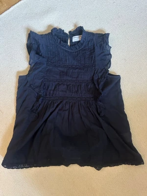 Mörkblå blus med volang från Holly & Whyte - Supersöt mörkblå blus från Holly & Whyte med volangkrage och dekorativa spetsdetaljer. Blusen är ärmlös.