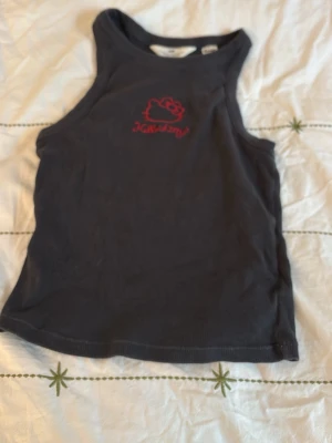 Svart Hello Kitty linnetopp - Säljer en svart linnetopp från Hello Kitty med ett rött broderat Hello Kitty-motiv och text på bröstet. Toppen är ärmlös och har en enkel, rak passform. Perfekt för dig som gillar söta detaljer och vill ha en bekväm topp till sommaren.