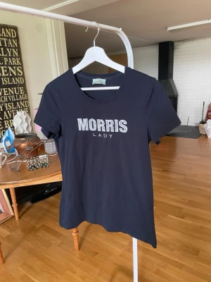 Svart Morris Lady t-shirt - Snygg svart t-shirt från Morris Lady med korta ärmar och rund hals. Framtill står det 'MORRIS LADY' där bokstäverna har ett blommigt mönster i blått och gult med vit kant. Perfekt till jeans eller kjol för en trendig look.