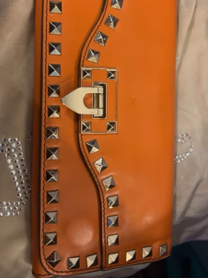 Orange plånbok Valentino Garavani - Snygg orange plånbok från Valentino Garavani i läder med guldfärgade nitar och dekorativt spänne framtill. Plånboken har en rektangulär form och en metallbricka med logga på baksidan. Perfekt accessoar för dig som gillar statement-detaljer.