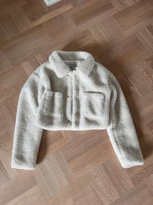 Vit/ljusbeige teddyjacka från NA-KD x Ivana - Säljer en kort, vit teddyjacka från NA-KD x Ivana Santacruz i storlek 36. Jackan har två stora bröstfickor, krage och stängs med knappar framtill. Supermjuk och fluffig i materialet, perfekt för att hålla värmen under kyliga dagar.