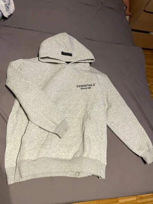 Grå Essentials Fear of God hoodie - Snygg grå hoodie från Essentials Fear of God med svart logga på bröstet. Klassisk passform med känguruficka och ribbade muddar. Tillverkad i mjuk bomullsblandning, perfekt för en avslappnad streetwear-look.