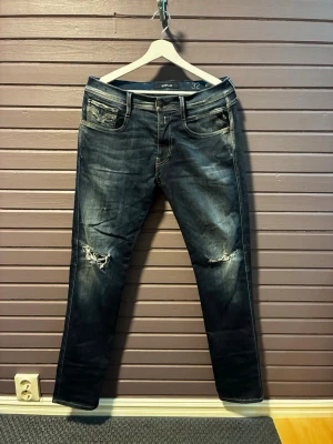 Mörkblå jeans med slitningar, Replay - Säljer ett par mörkblå jeans från Replay med slitningar på båda knäna. Klassisk femficksmodell med normal passform och snygg tvätt. Perfekta för dig som gillar en avslappnad men trendig stil.