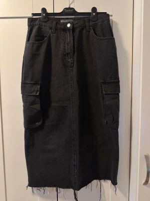 Svart jeanskjol med cargofickor - Säljer en svart lång jeanskjol med cargofickor på sidorna och rå kant nertill. Kjolen har klassisk femficksdesign, bälteshällor och stängs med knapp och dragkedja framtill. Perfekt för en cool och avslappnad stil. Köpt från London