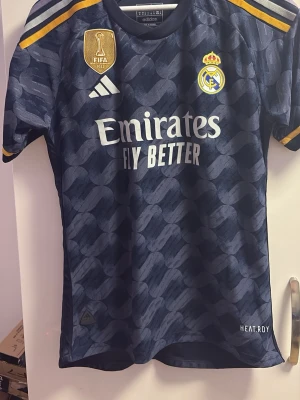 Real Madrid bortatröja Adidas blå - Snygg Real Madrid bortatröja i mörkblått med mönster, gula detaljer och klubbmärke på bröstet. Adidas-logga, FIFA-märke och sponsortryck framtill. Tillverkad i funktionsmaterial HEAT.RDY för bästa komfort vid träning eller match. Bellingham på ryggen