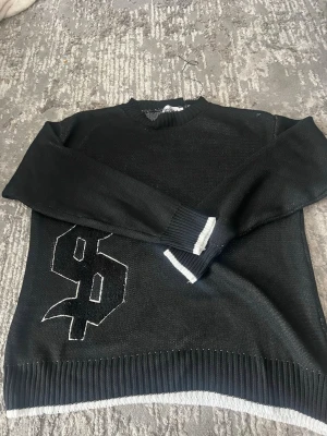 Svart stickad  - Svart stickad tröja från Noon Studio med stor vit broderad siffra på framsidan. Tröjan har ribbstickade muddar och nederkant med vita detaljer. Klassisk rund halsringning och avslappnad passform.