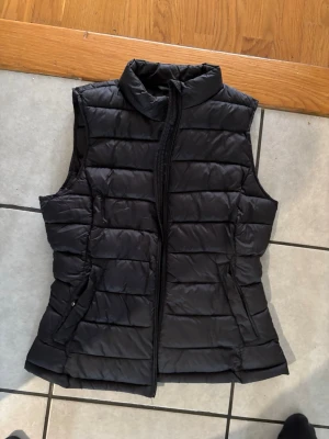 Svart quiltad dunväst med dragkedja - Stilren svart dunväst med quiltad design och hög krage. Västen har två fickor med dragkedja och stängs med en hel dragkedja framtill. Perfekt för lager-på-lager och enkel att matcha till olika outfits.