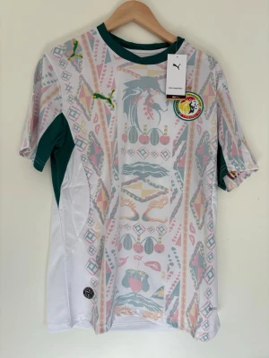 Senegal fotbollströja Puma vit/grön - Säljer en officiell Senegal fotbollströja från Puma i vitt med gröna detaljer och färgglatt mönster framtill. Tröjan har korta ärmar, rund hals och Senegal-emblem på bröstet. Tillverkad i lätt och ventilerande material, perfekt för träning eller match.