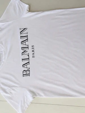 Vit Balmain Paris t-shirt - Säljer en vit t-shirt från Balmain Paris med klassiskt svart logotryck på bröstet. T-shirten har rund hals och korta ärmar, perfekt för dig som vill ha en clean och stilren look. Pris 499
