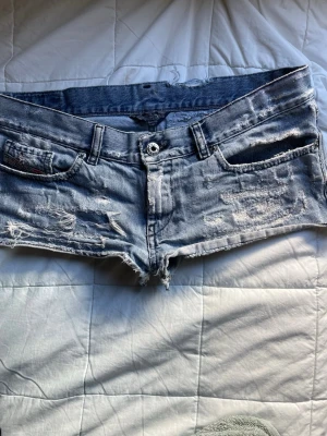 Lågmidjade slitna jeansshorts - Säljer ett par ljusblå lågmidjade jeansshorts från diesel med slitna detaljer och råa kanter. Klassisk femficksmodell med knappgylf och coolt vintage-vibe. Perfekta för varma dagar och en avslappnad stil.