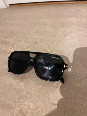 Svarta Tom Ford pilotbågar solglasögon - Snygga svarta solglasögon från Tom Ford med klassisk pilotform och mörka glas. Bågarna har gulddetaljer vid gångjärnen och logga på linsen. Perfekta för dig som vill ha en stilren och exklusiv look.