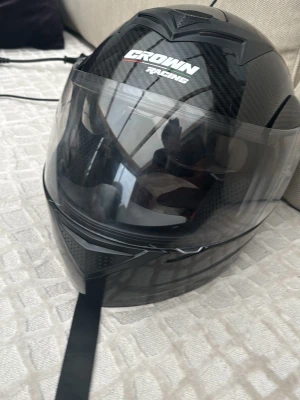 Svart motorcykelhjälm  från Crown Racing - Snygg svart hjälm från Crown Racing med kolfiberlook och klart visir. Hjälmen har en sportig design med ventilationsdetaljer och justerbart hakband. Perfekt för dig som vill ha en cool och säker look på vägarna. Den har dock inget spänne på så du får gärna köpa en men annars är den i bra skick 
