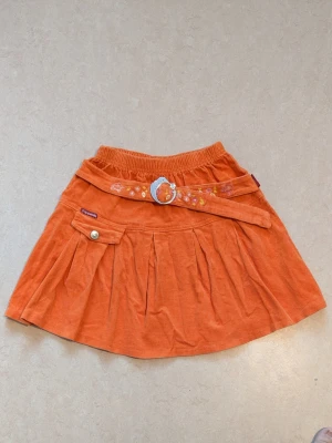 Orange manchesterkjol med bälte - Superfin orange kortkjol i manchester. Kjolen har ett dekorativt bälte med broderade blommor och en söt ficka med knapp framtill. Perfekt för dig som vill sticka ut med färg och detaljer!