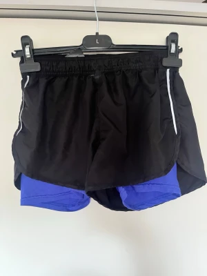 Svarta löparshorts med blå innershorts - Säljer ett par svarta löparshorts från Crivit med blå innershorts. Shortsen har elastisk midja och vita detaljer längs sidorna. Tillverkade i 100% polyester vilket gör dem lätta och snabbtorkande. Perfekta för löpning eller träning. Superfina nya endast tvättade 