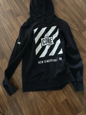 Svart hoodie Jack & Jones CORE - Snygg svart hoodie från Jack & Jones med stor vit CORE-logga och grafiskt tryck framtill. Texten 'NEW DIMENSIONS | 90' finns under trycket och siffran 90 på ärmen. Mjuk insida och klassisk huva med dragsko. Perfekt för en avslappnad streetwear-look.