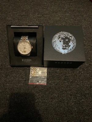 Versus Versace silver/guld klocka - Snygg klocka från Versus Versace i silver och guld med klassisk rund urtavla och detaljerat mönster. Klockan har metallarmband och ett lejonmotiv på urtavlan. Kommer i originalask med extra länkar.
