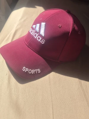 Vinröd Adidas keps med brodyr - Snygg vinröd keps från Adidas med vit broderad logga och texten 'SPORTS' framtill. Klassisk böjd skärm och justerbart spänne bak i metall. Tillverkad i slitstark bomull, perfekt för en sportig och avslappnad stil.
