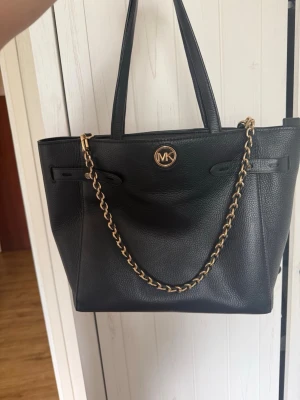 Svart Michael Kors handväska i läder - Snygg svart handväska från Michael Kors i strukturerat läder. Väskan har guldfärgade detaljer, inklusive en kedja och MK-logga framtill. Två långa handtag gör den enkel att bära på axeln. Perfekt för dig som gillar klassisk och stilren design. Ny pris 3000kr