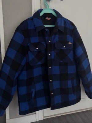 Blå och svart overshirt från Dickies - Snygg overshirt från Dickies i blått och svart rutigt mönster. Jackan har två bröstfickor med knappar, två sidofickor och klassisk krage. Perfekt lager på lager-plagg med avslappnad passform och cool streetwear-vibe. Knappt använd bra skick är ett litet bränn hål på kragen bak se sista bild!
