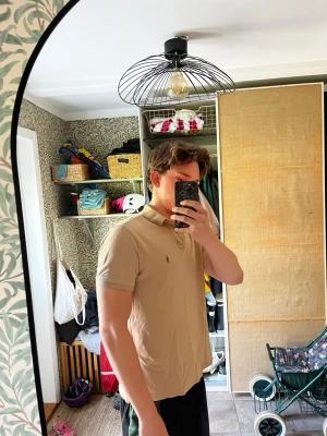 Beige pikétröja från Ralph Lauren - Snygg beige pikétröja från Ralph Lauren med klassisk krage och knappar framtill. Tröjan har korta ärmar och den ikoniska loggan broderad på bröstet. Perfekt till jeans eller shorts för en clean och stilren look.