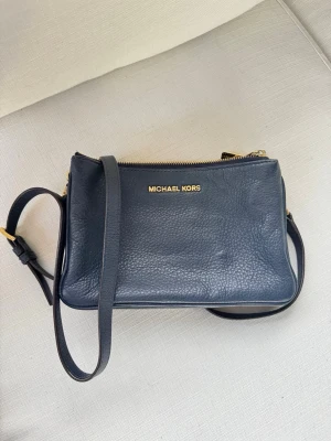 Mörkblå axelväska från Michael Kors - Snygg mörkblå axelväska från Michael Kors i äkta läder med guldfärgade detaljer och tre separata fack med dragkedja. Justerbar axelrem och stilren design med logga framtill. Perfekt för att hålla ordning på dina grejer.