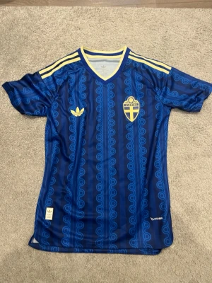 Blå Sverige fotbollströja Adidas - Säljer en blå fotbollströja från Adidas med Sveriges landslagsemblem på bröstet. Tröjan har gula detaljer, klassiska Adidas-ränder på axlarna och ett unikt mönster i olika blå nyanser. Tillverkad i lätt och ventilerande material, perfekt för träning eller match.
