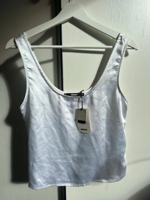 Vit satinlinne från Bik Bok - Snyggt vitt linne från Bik Bok i glansigt satinmaterial. Linnet har breda axelband och en djup rundad urringning både fram och bak. Perfekt att styla med jeans eller kjol för en trendig look.
