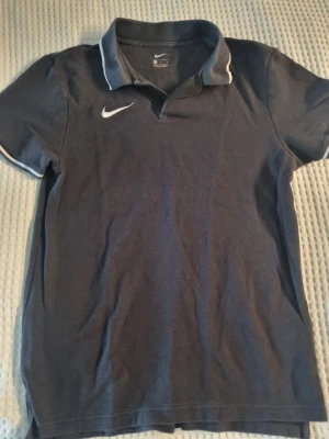 Pikétröja från Nike - Snygg pikétröja från Nike med vit logga på bröstet och vita detaljer på krage och ärmslut. Klassisk pikékrage och korta ärmar. Perfekt för en sportig och avslappnad stil.