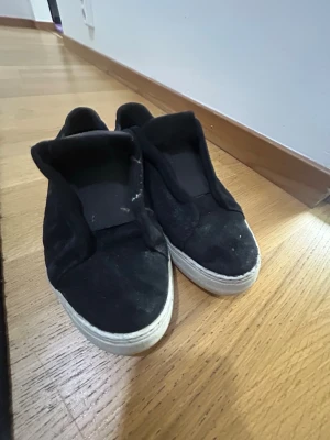 Svarta slip-on sneakers - Svarta slip-on sneakers med vit sula och ovandel i mockaliknande material. Skorna har en enkel och stilren design utan snörning, vilket gör dem lätta att ta på och av. Passar perfekt till en avslappnad streetwear-look.