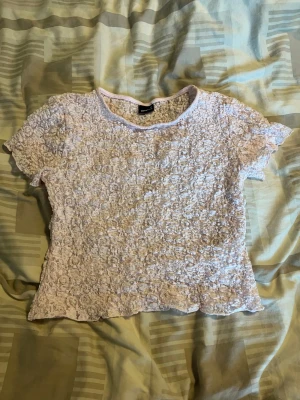Ljusrosa spetstopp - Supersöt ljusrosa topp med korta ärmar och heltäckande blommönster i spets. Toppen har en rund halsringning och är lätt figursydd för en feminin look. Perfekt att styla med jeans eller kjol