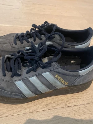 Adidas Spezial blå sneakers - Säljer ett par Adidas Spezial sneakers i grå mocka med ljusblåa klassiska tre ränder på sidan och guldfärgad Spezial-text. Skorna har mörkblå snören och sula i gummi. Perfekta för dig som gillar retrostil och streetwear.