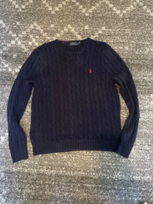 Mörkblå kabelstickad tröja Polo Ralph Lauren - Säljer en mörkblå kabelstickad tröja från Polo Ralph Lauren med rund halsringning och den klassiska röda logotypen broderad på bröstet. Tröjan har långa ärmar och ribbade muddar vid ärmslut och nederkant. Perfekt för dig som gillar stilrena och tidlösa plagg.